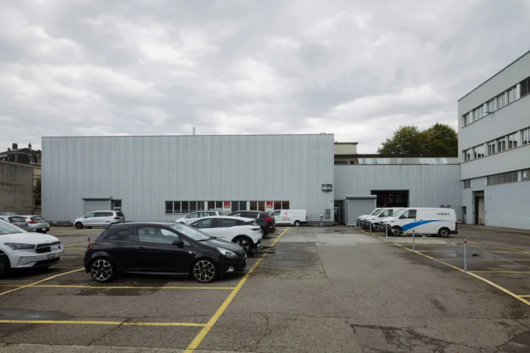 Parkplatzfläche im Vitus-Areal Winterthur mit geparkten Autos und einer Industriehalle im Hintergrund.