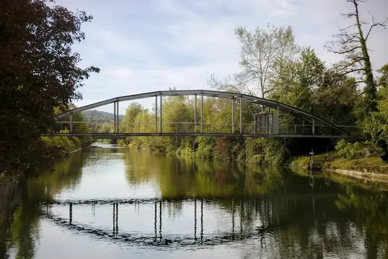Stahlbrücke über die Töss beim Vitus-Areal Winterthur mit Spiegelung im ruhigen Fluss und umgebender Ufervegetation.