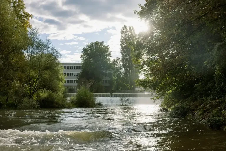 Blick auf die Töss beim Vitus-Areal in Winterthur mit Flusslauf, Ufervegetation und dahinterliegendem Industriegebäude.