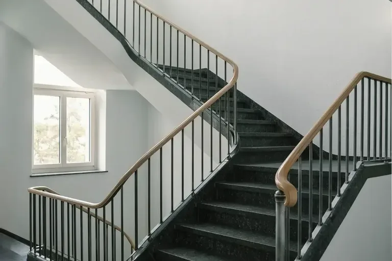 Treppe im Vitus-Areal Winterthur mit dunklen Stufen, Metallgeländer und Holzhandlauf in einem hellen Treppenhaus.